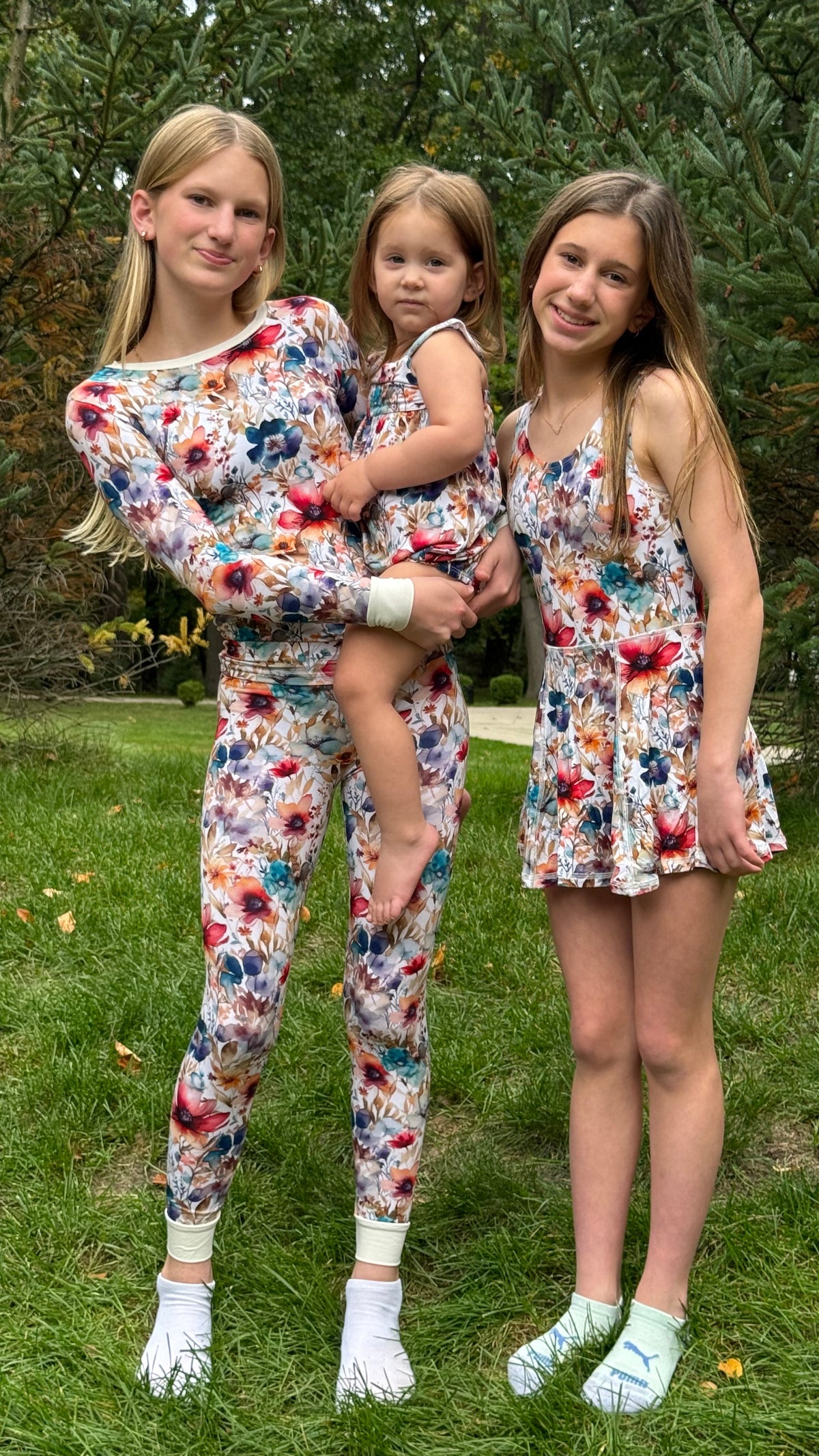 Three girls wearing matching floral print matching sets pose together outdoors on a grassy lawn with trees in the background. The set includes a long-sleeve top and leggings on the left girl, a sleeveless mini dress on the right girl, and a matching set on the toddler held in the center.